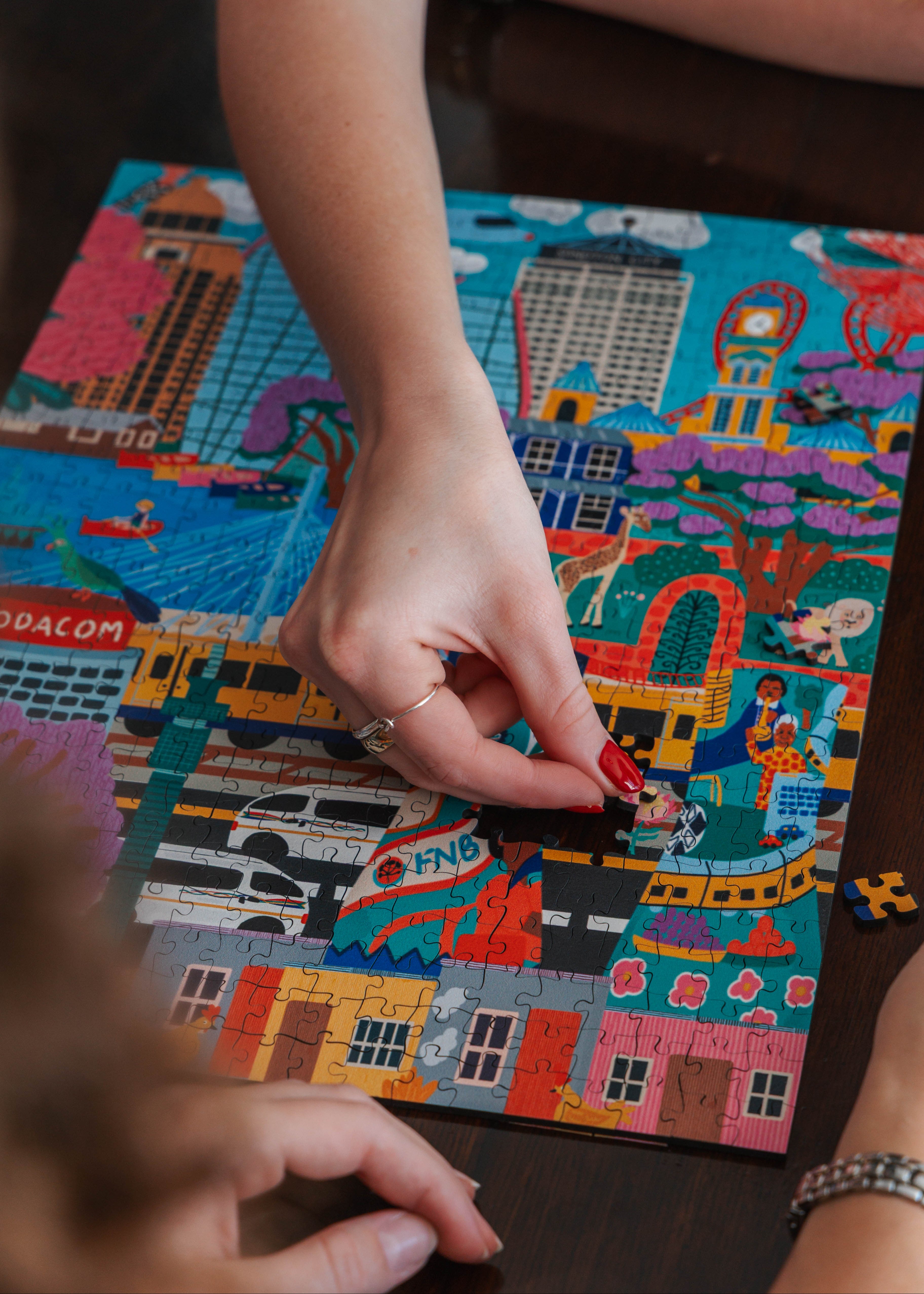 Person assembling a colorful puzzle with various images and text on a wooden table. Johannesburg by Kelly McChesney Puzzle