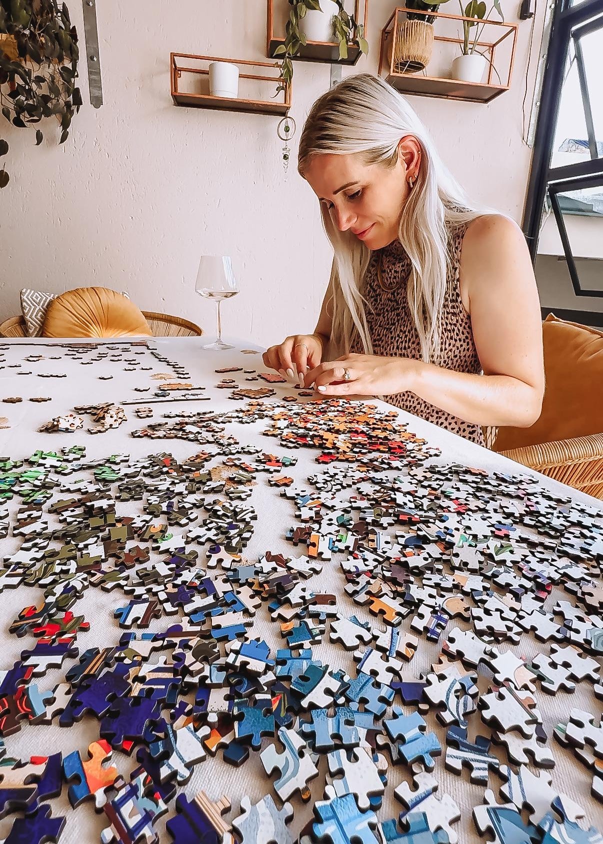 Woman working on a large puzzle with scattered pieces in a room with plants and shelves.