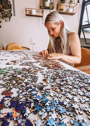 Woman working on a large puzzle with scattered pieces in a room with plants and shelves.