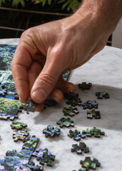 Hand placing puzzle pieces on a partially completed puzzle on a marble surface.