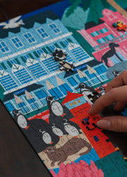Puzzle with cartoon characters and colorful buildings, hand placing a piece. Simon's Town Penguins