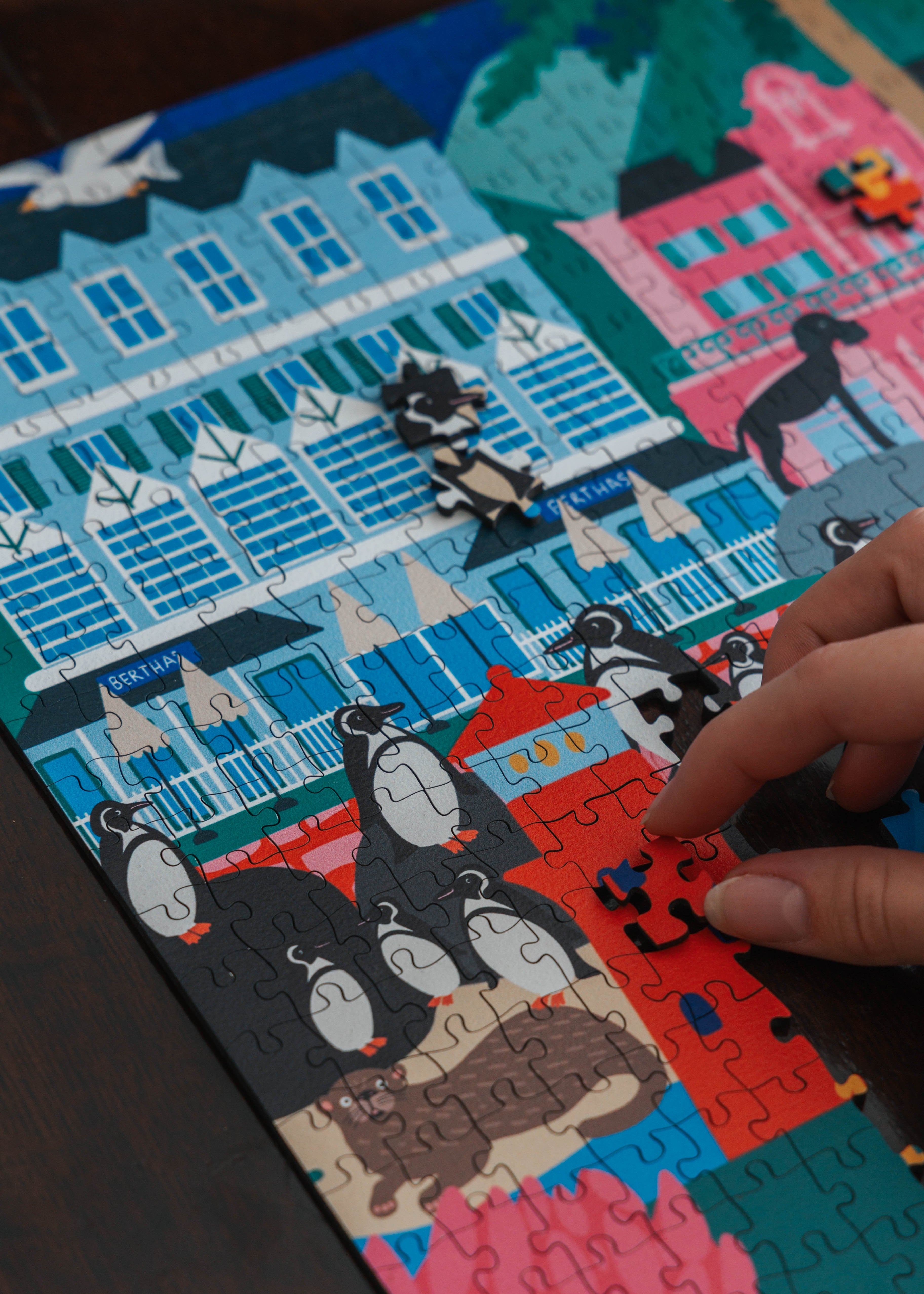 Puzzle with cartoon characters and colorful buildings, hand placing a piece. Simon's Town Penguins