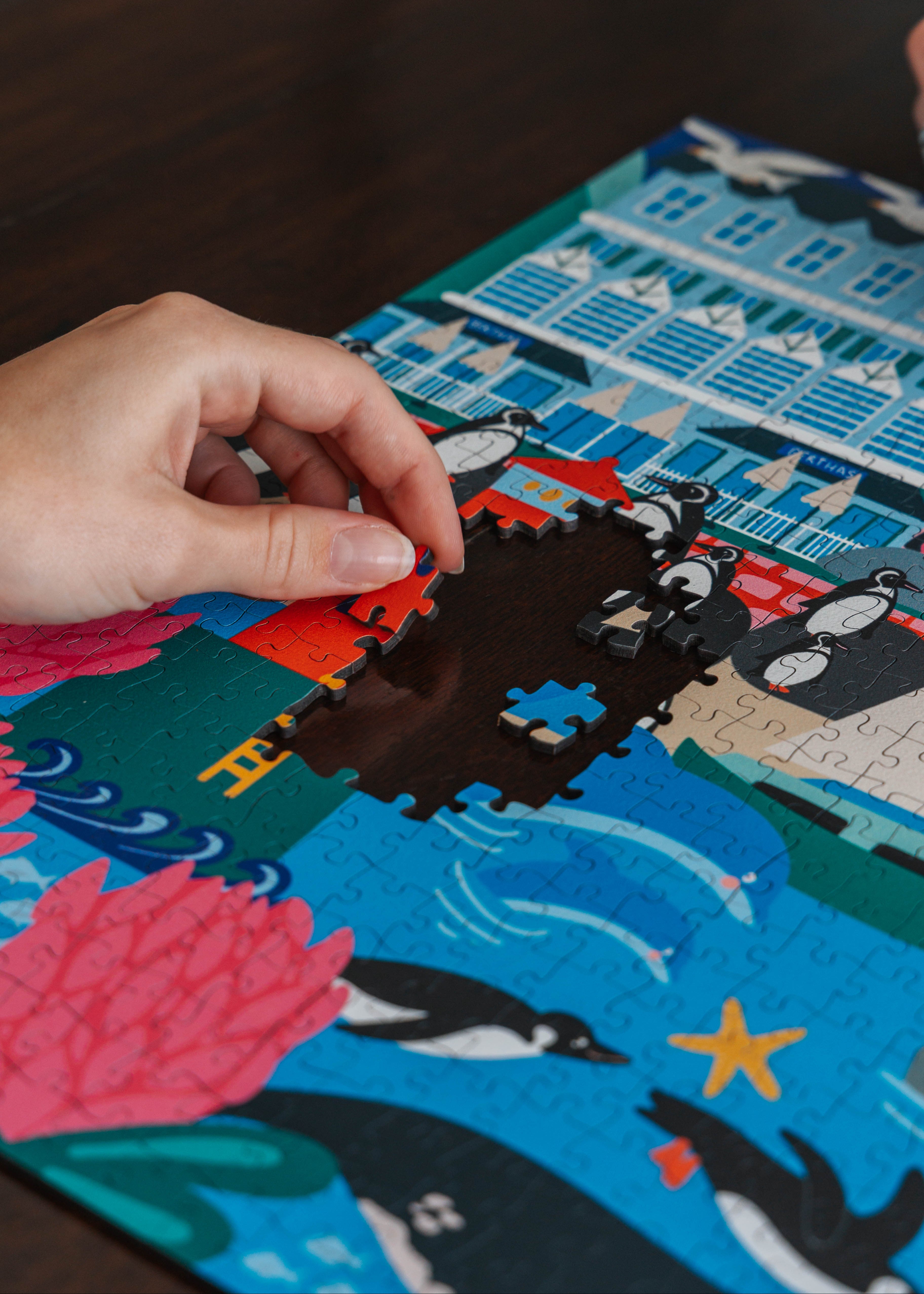 Hand placing a puzzle piece on a colorful puzzle with penguin design. Simon's Town Penguins